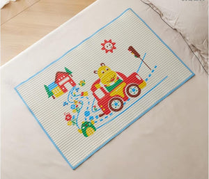 A rectangular white textured mat with a light blue border, featuring a colorful cartoon fire truck scene with a yellow bear, red truck, blue hose, and small house, placed on a light beige bed or cushion.