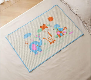 A rectangular baby mat with a textured waffle pattern, featuring a colorful cartoon design of a blue elephant, orange giraffe, and sun, bordered in light blue, shown laid out on a white bed.