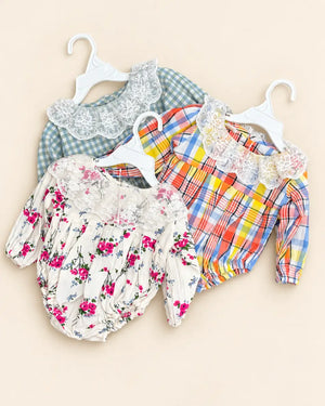 Three baby long-sleeve bloomers on white plastic hangers, displayed against a light beige background. The frontmost pair is cream with a pink and red floral print, the middle pair is yellow, orange, and blue plaid with a white lace collar, and the back pair is green and blue gingham with a white lace collar.