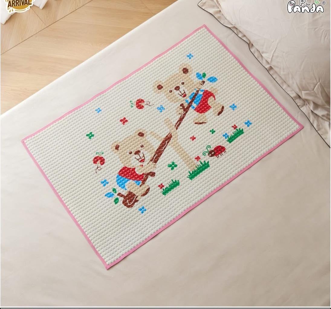 A rectangular woven mat with a pink border and a cartoon teddy bear design, placed on a bed. The mat features a white textured center with a pink border, a brown teddy bear holding a guitar, and small colorful embroidered details.
