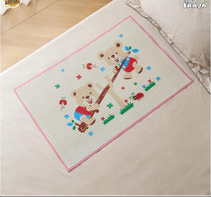 A rectangular woven mat with a pink border and a cartoon teddy bear design, placed on a bed. The mat features a white textured center with a pink border, a brown teddy bear holding a guitar, and small colorful embroidered details.